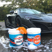 Two buckets labeled 'Rinse' and 'Wash' in front of a black sports car on a wet road.